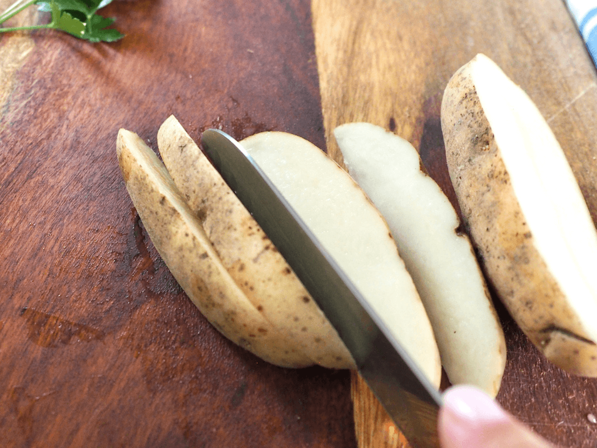 Knife slicing russet potatoes on cutting board into wedges.