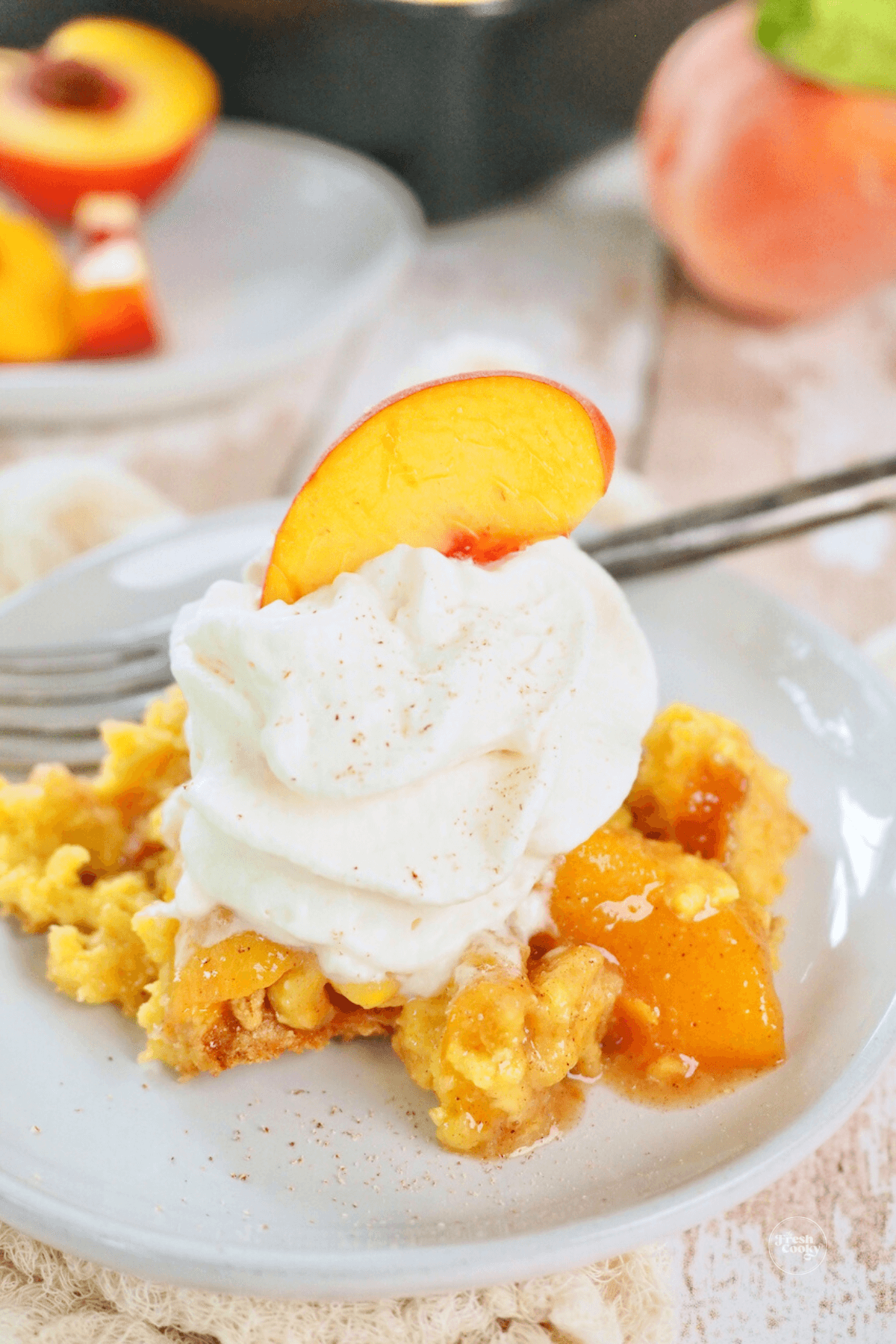 Slice of peach cobbler on plate with bourbon whipped cream and topped with a fresh slice of peach.