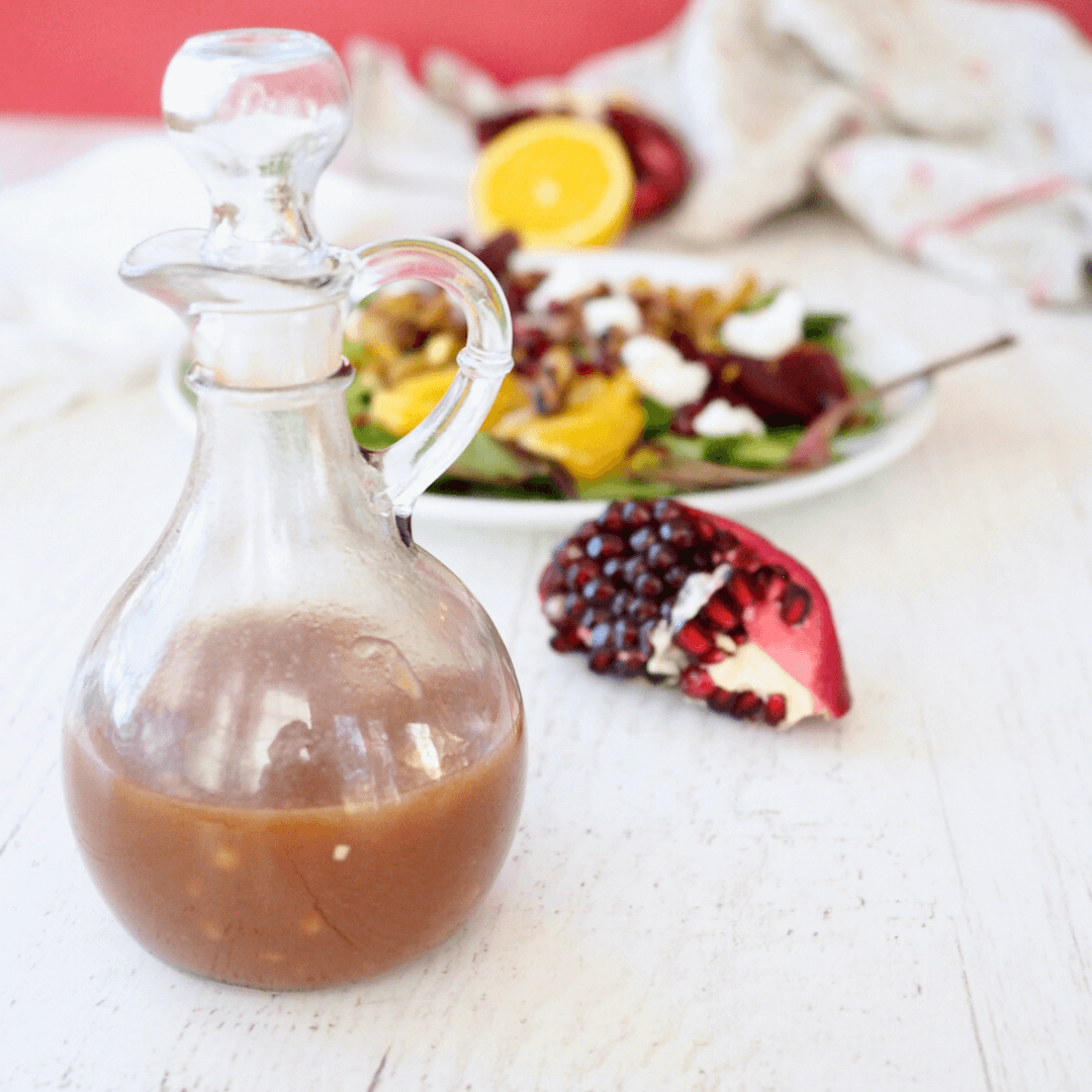 Pomegranate dressing in cruet with salad behind and quarter of pomegranate on counter.