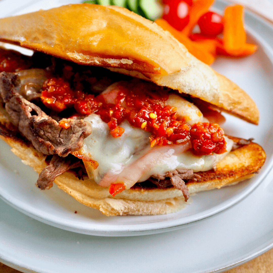 Steak bomb sandwich on white plate with veggies behind.