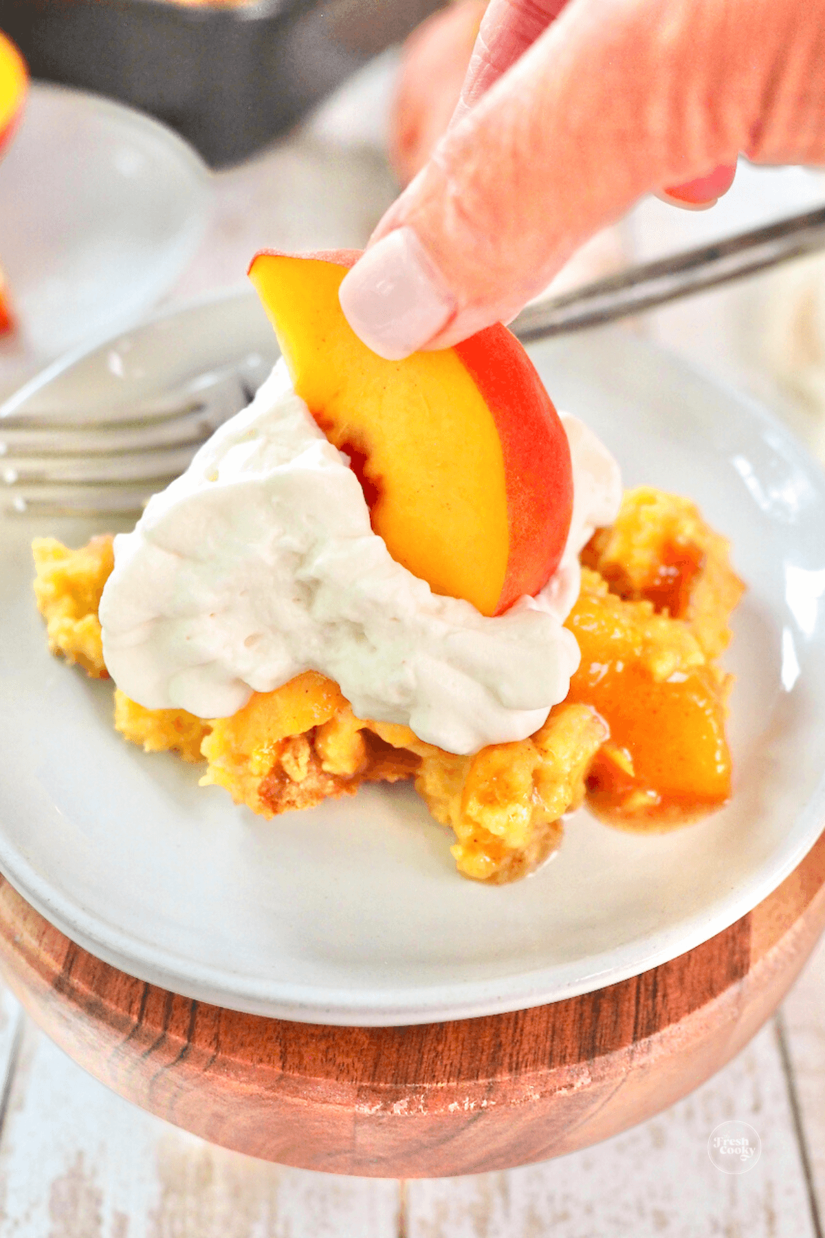 Hand adding a sliced peach to the top of serving of peach cobbler with whipped cream.