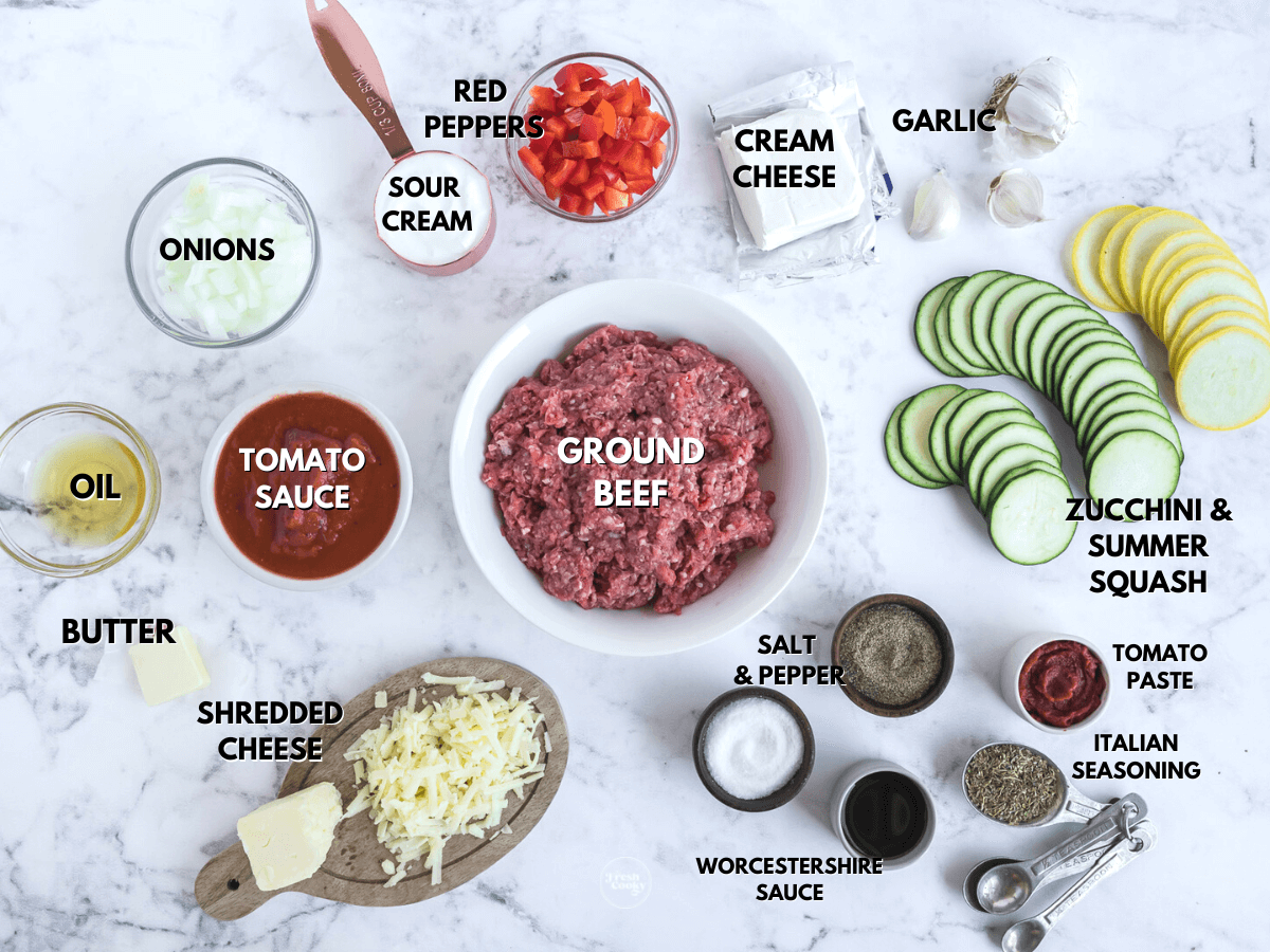 Labeled ingredients for ground beef zucchini casserole L-R onions, bell peppers, sour cream, cream cheese, garlic, zucchini, yellow squash, tomato paste, Italian seasoning, ground beef, cheese, tomato sauce, butter, oil.