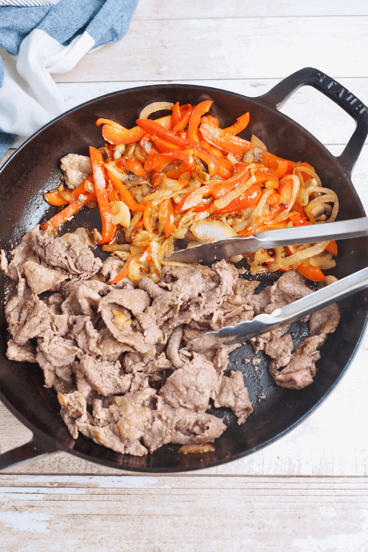 Browning shaved steak in skillet with peppers and onions on side. 