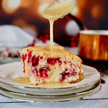 Cranberry Christmas cake slice on a plate while a spoon drizzles vanilla butter sauce on top.