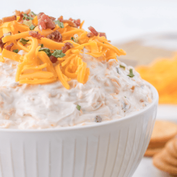 Cowboy crack dip recipe in pretty white bowl with crackers and shredded cheese in background.