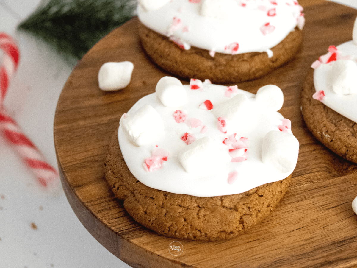 Hot cocoa cookies frosted with marshmallow fluff and crushed candy canes in a wooden pedestal.