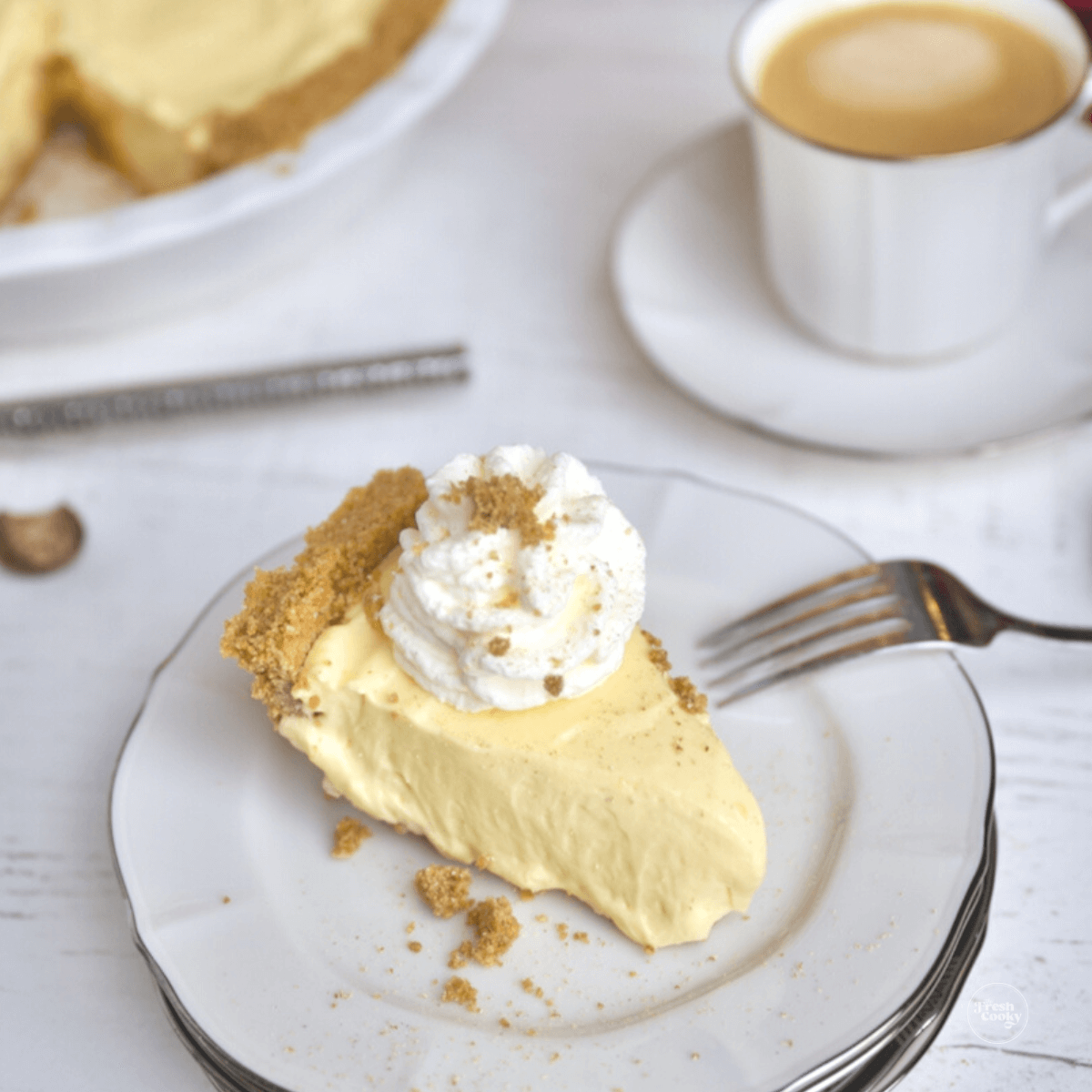 Slice of no bake eggnog pie on plate and topped with whipped cream with coffee in background.