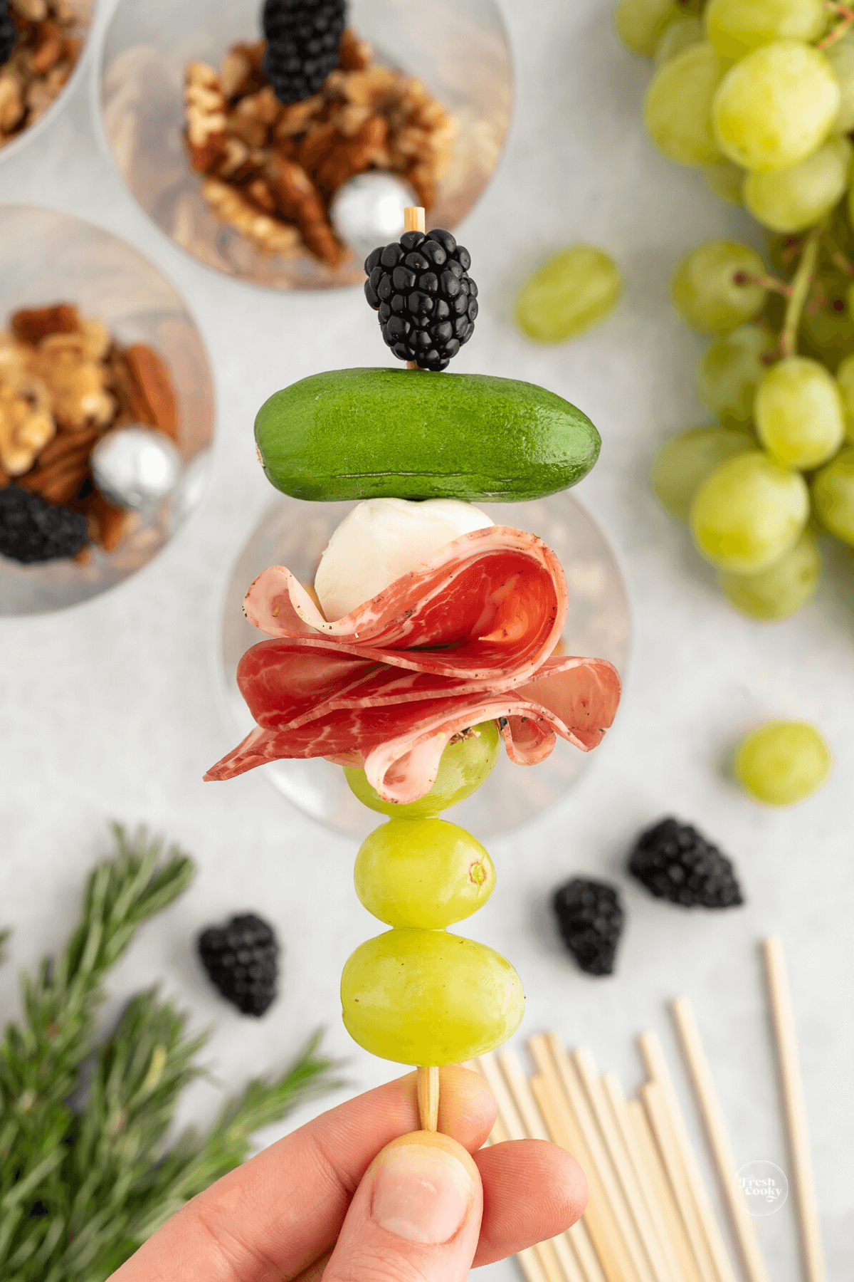 Hand holding skewer packed with grapes, salami, baby mozzarella, mini cucumber, blackberry.