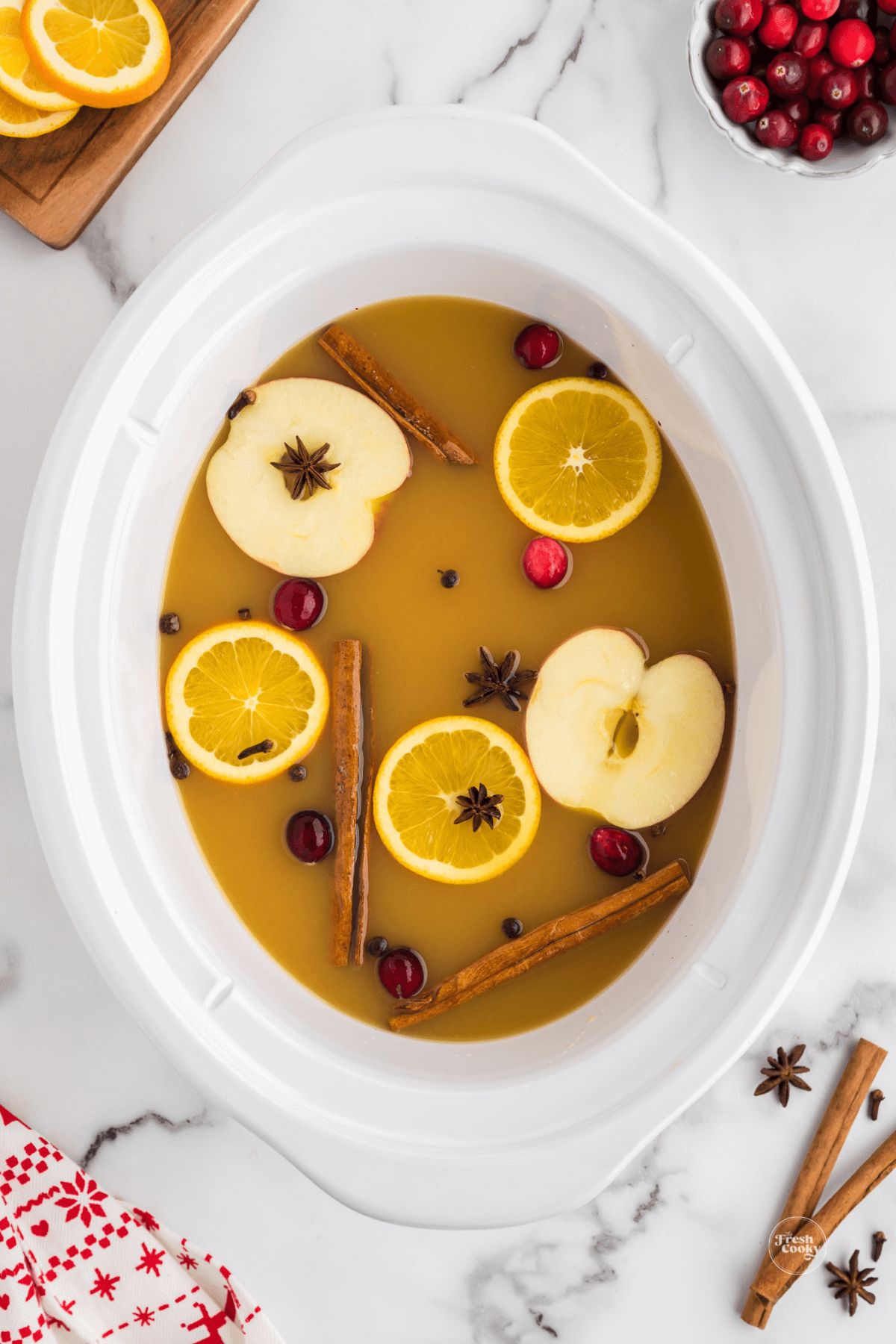 Hot mulled cider wassail in white crockpot with floating apple slices, oranges and spices.