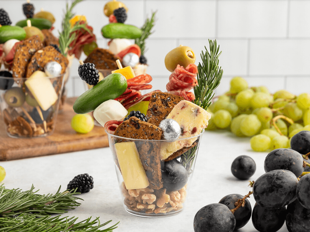 How to pack charcuterie board to go, showing cups on a board and charcuterie cup on counter with fresh herbs and grapes.