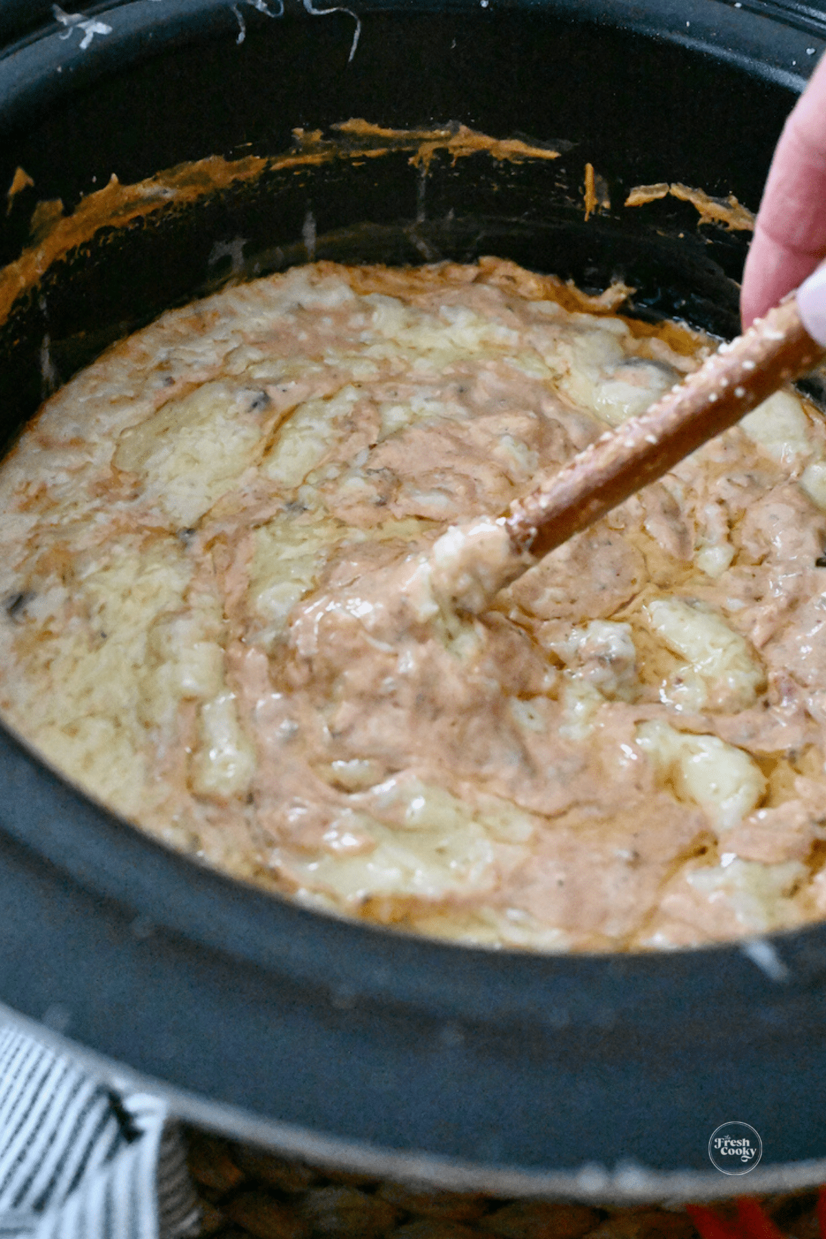 Hand dipping a pretzel rod into gooey chili cheese dip in crock pot.