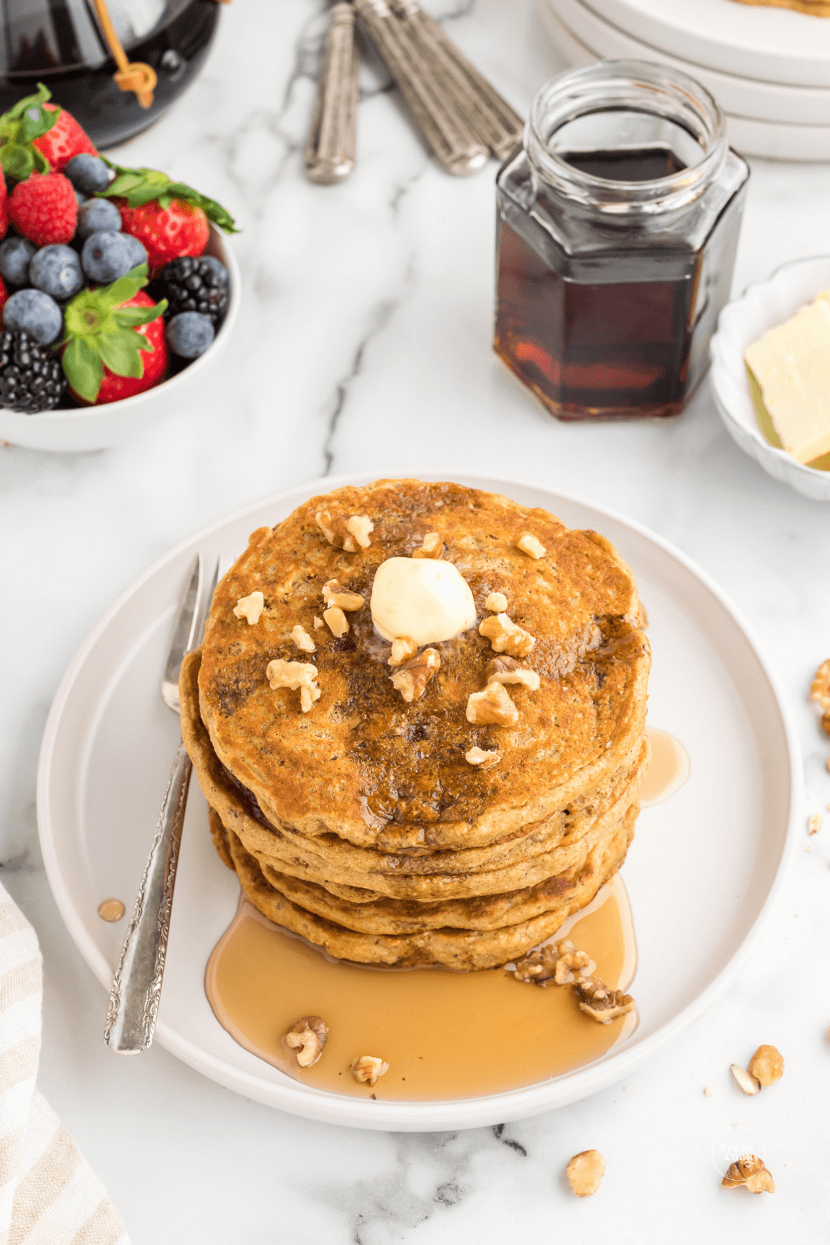 Stack of pancakes with soft butter and oozing maple syrup topped with walnuts with fresh berries in background and maple syrup.