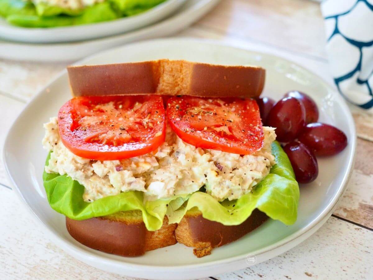 Southern Tuna Salad sandwich, with lettuce and thick juicy tomatoes with grapes, on a pretty white pottery plate.
