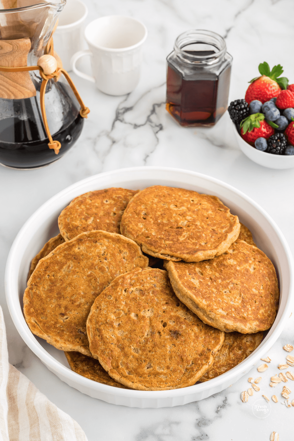 Warm pancakes ready for serving on platter with fresh fruit, maple syrup and coffee behind.