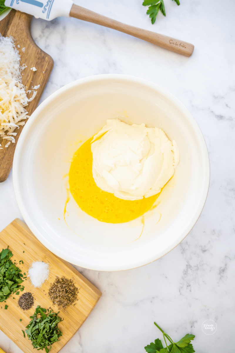 Beaten egg with ricotta cheese in bowl, spices nearby for stuffed shells. 