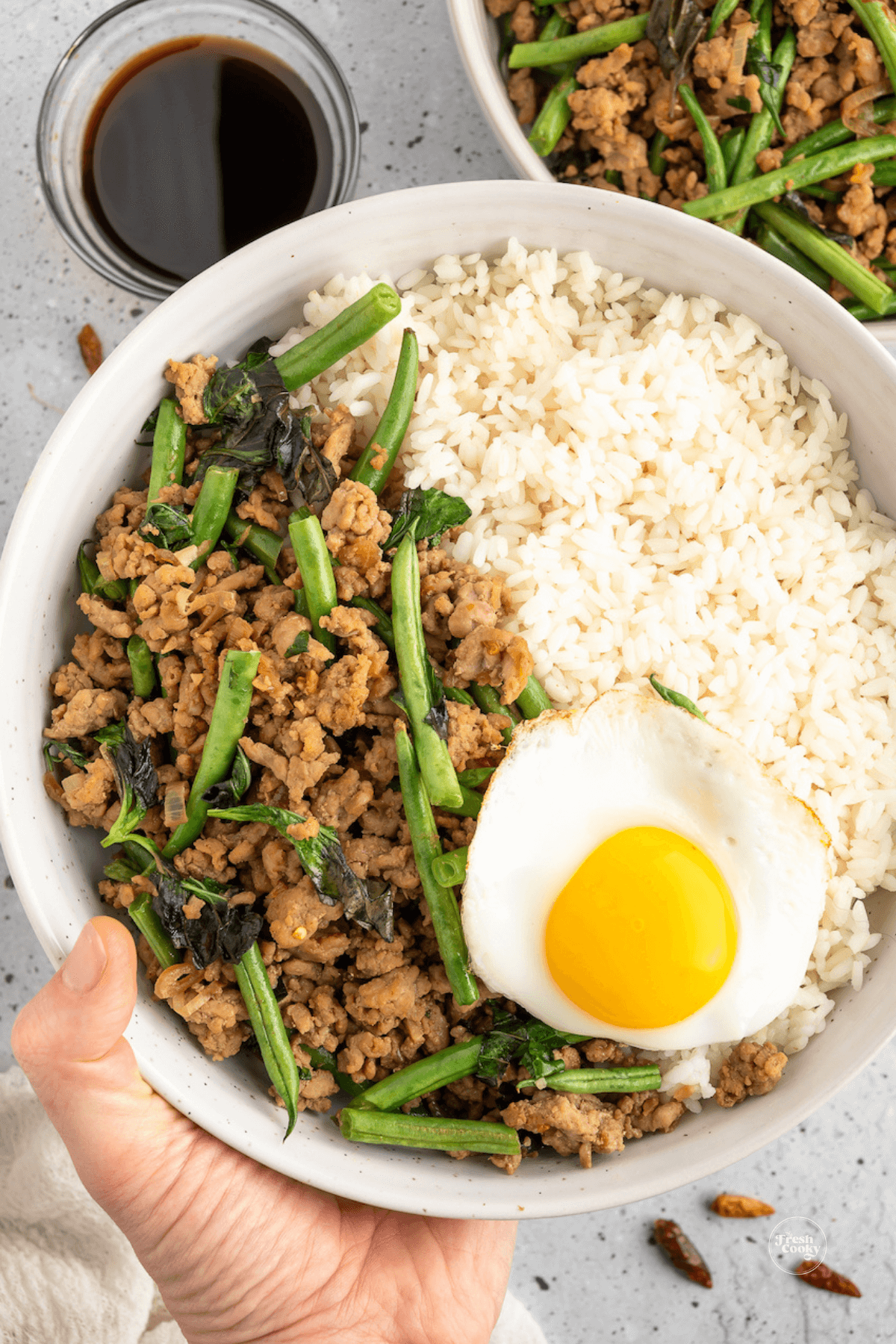 Hand holding bowl of ground pork stir fry with Thai fried egg and rice.