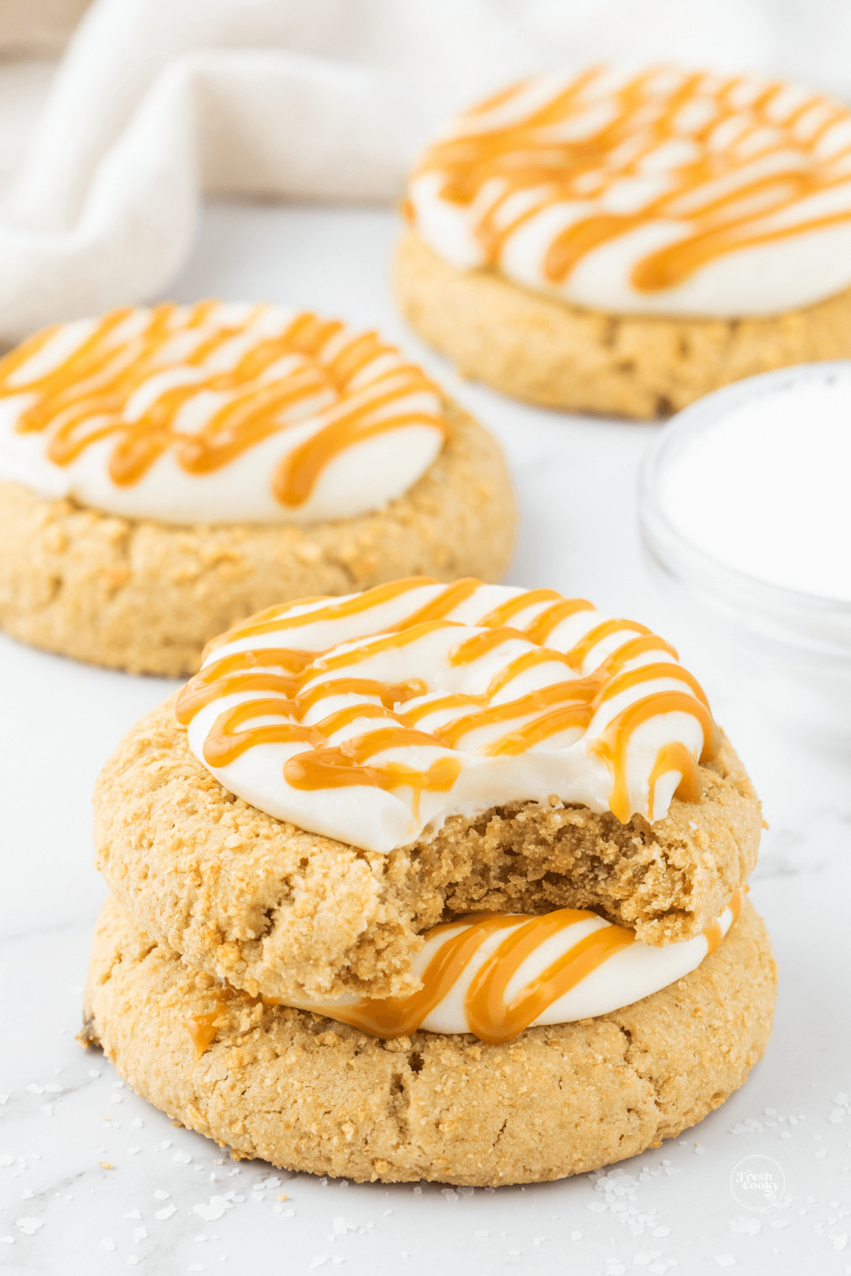 Salted caramel cheesecake cookies stacked one on another, with other cookies behind, frosted and drizzled with caramel.