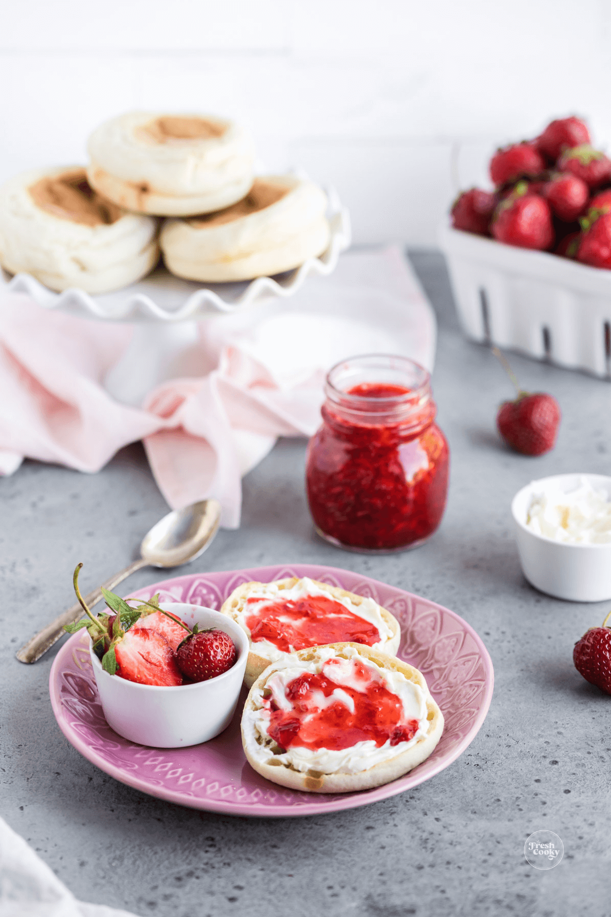 Strawberry Freezer jam recipe in a jar with plate of toasted english muffins topped with fresh jam and strawberries in background.