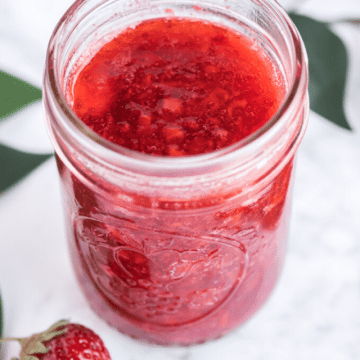 Strawberry Freezer Jam in a mason jar with a berry on the counter.