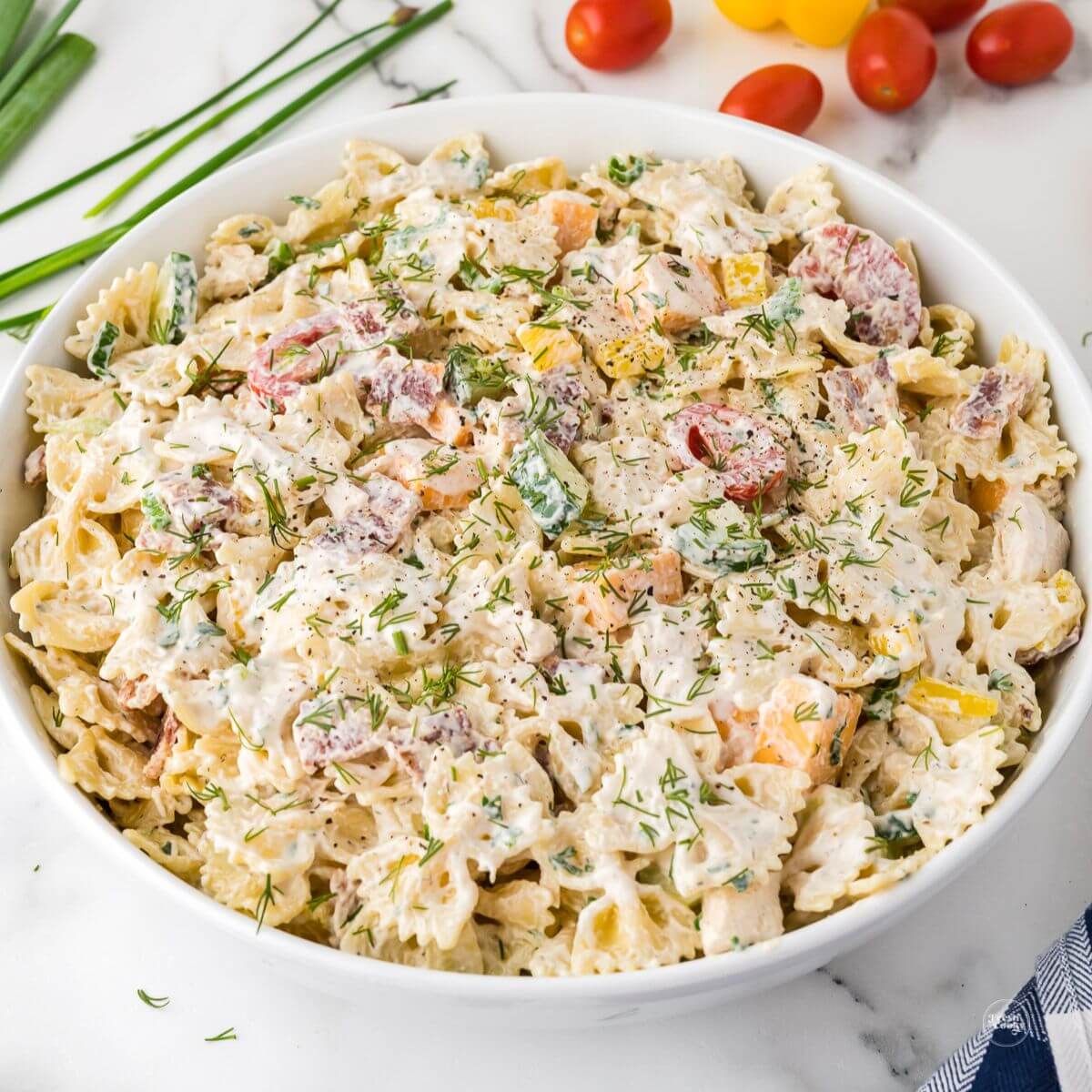 Chicken bacon ranch pasta salad in a large white bowl with fresh tomatoes and herbs behind.