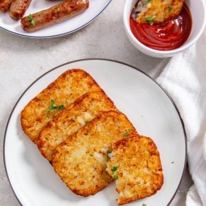 hash brown patties in air fryer on plate with ketchup and sausage links behind.