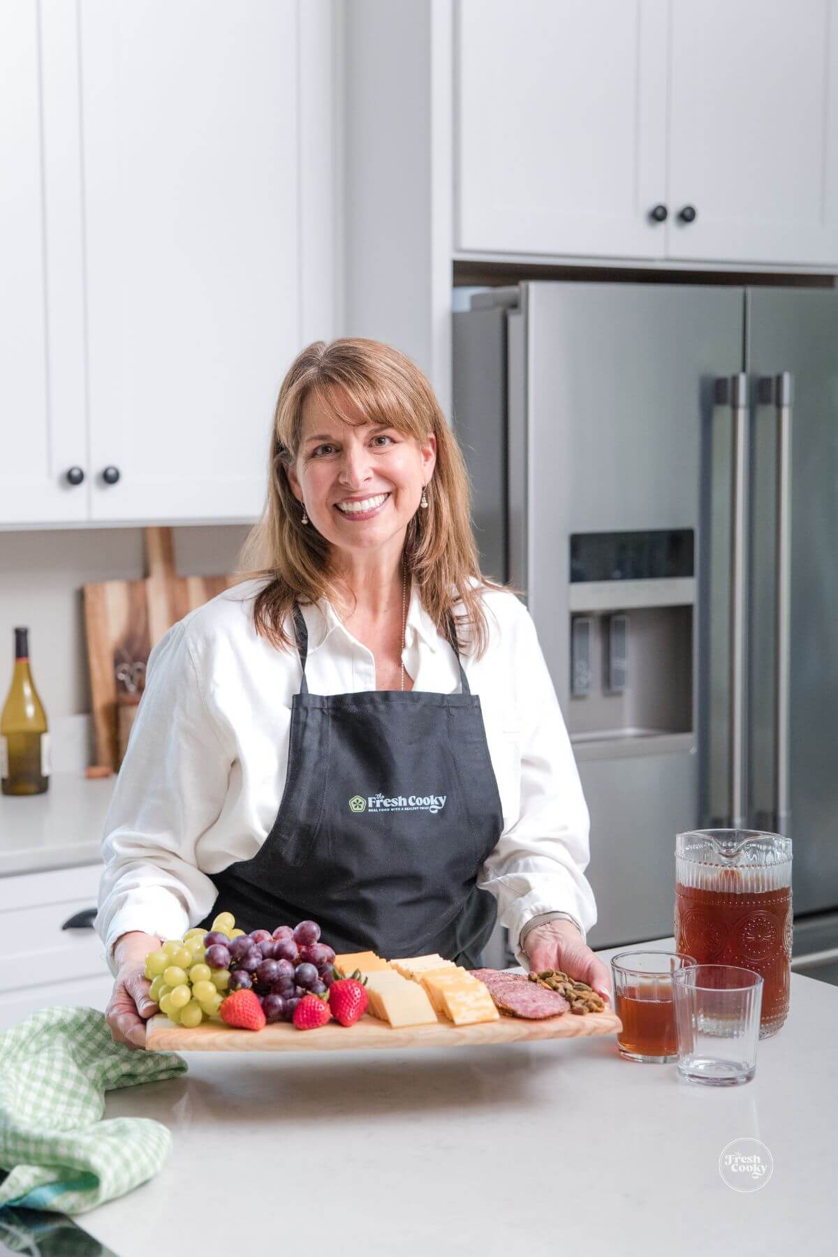 Welcome to The Fresh Cooky! Kathleen, holding a charcuterie board with a Fresh Cooky Apron on.