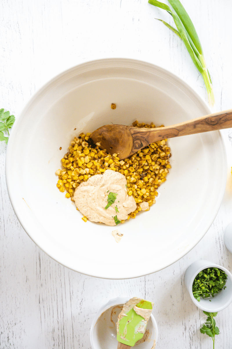 Adding elotes dressing to roasted corn in bowl. 