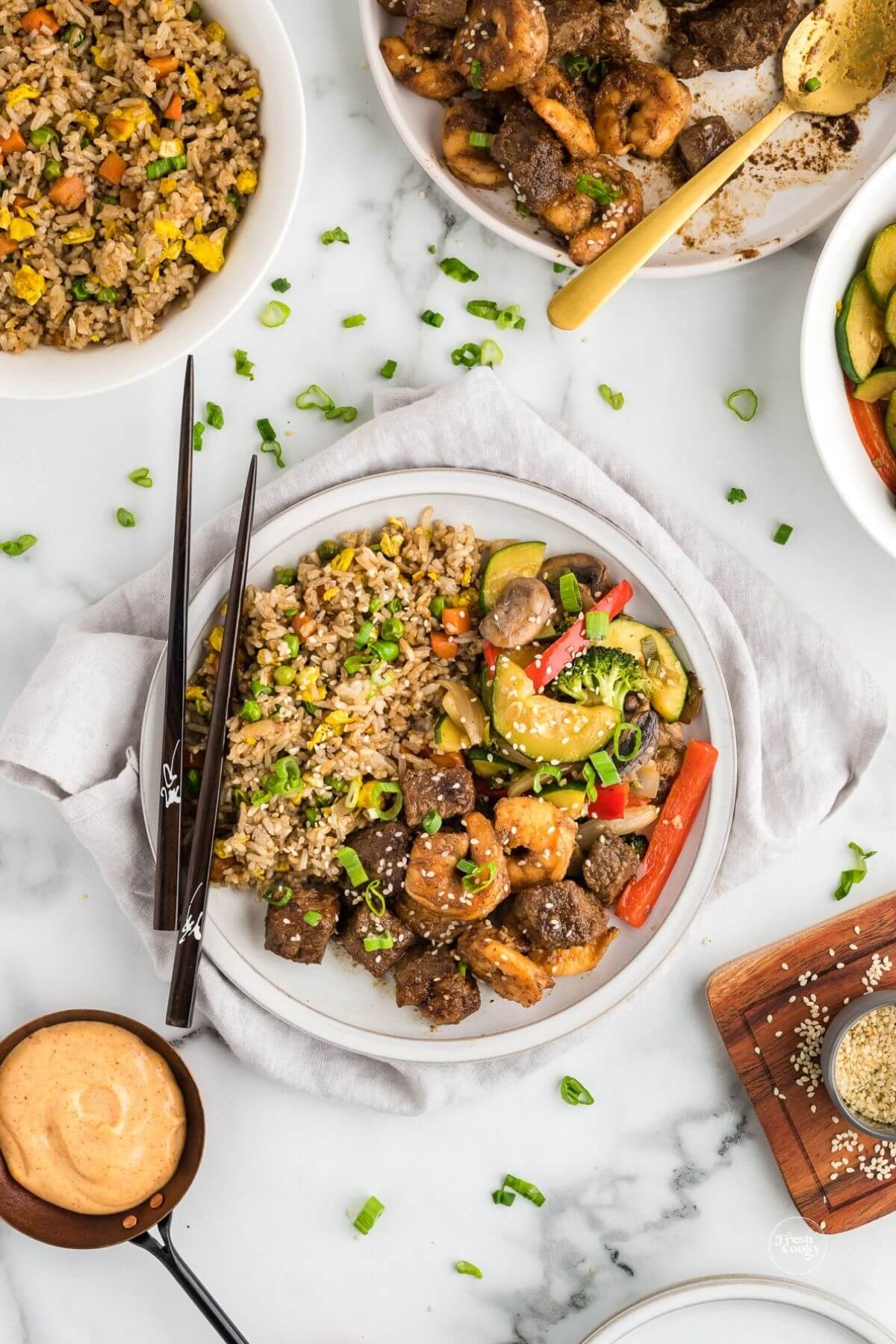 HIbachi steak and shrimp recipe plated with yum yum sauce and bowls of fried rice, hibachi veggies nearby.