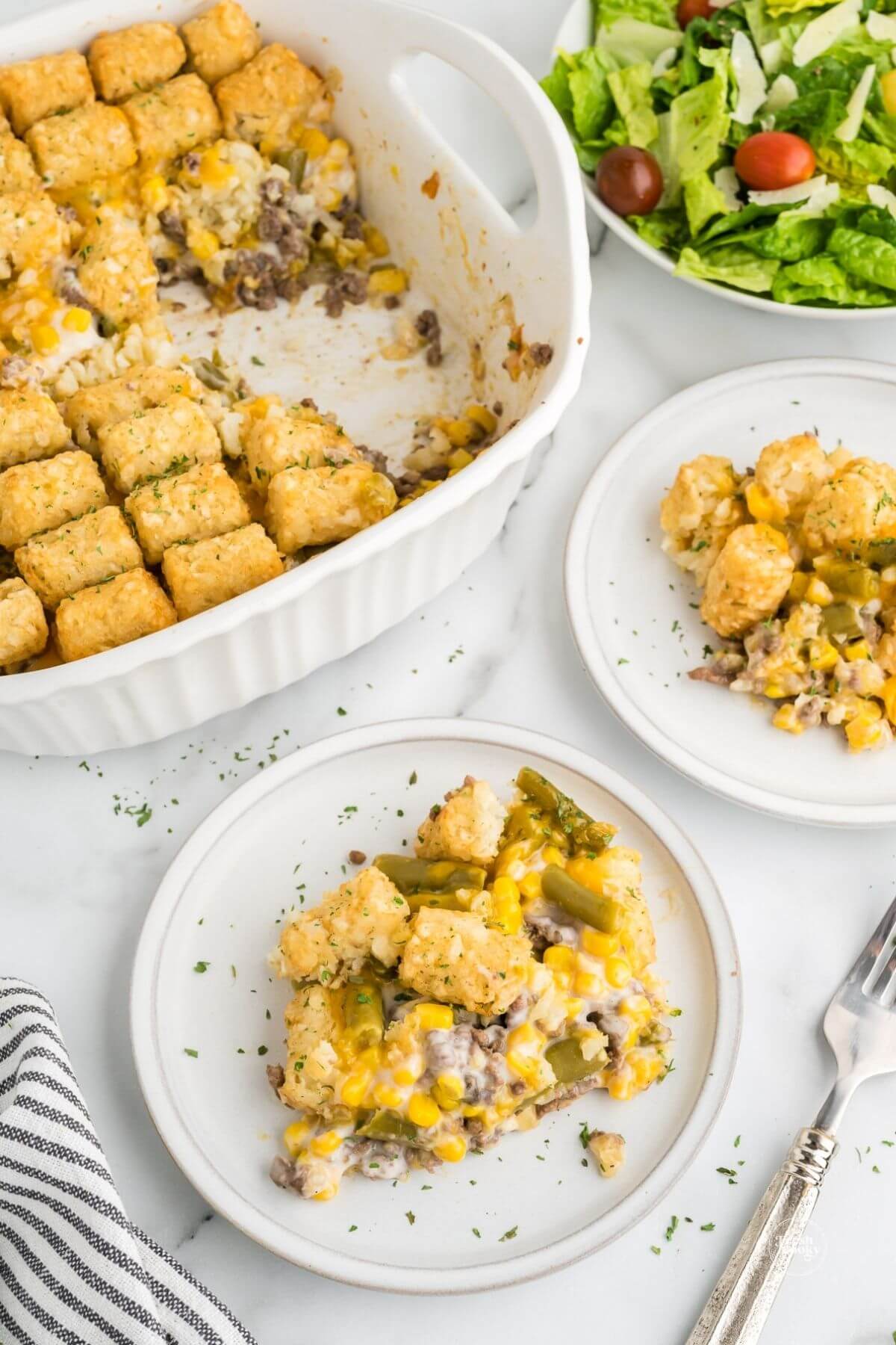 Two servings of cheesy hamburger tater tot casserole recipe on plate with casserole dish behind.