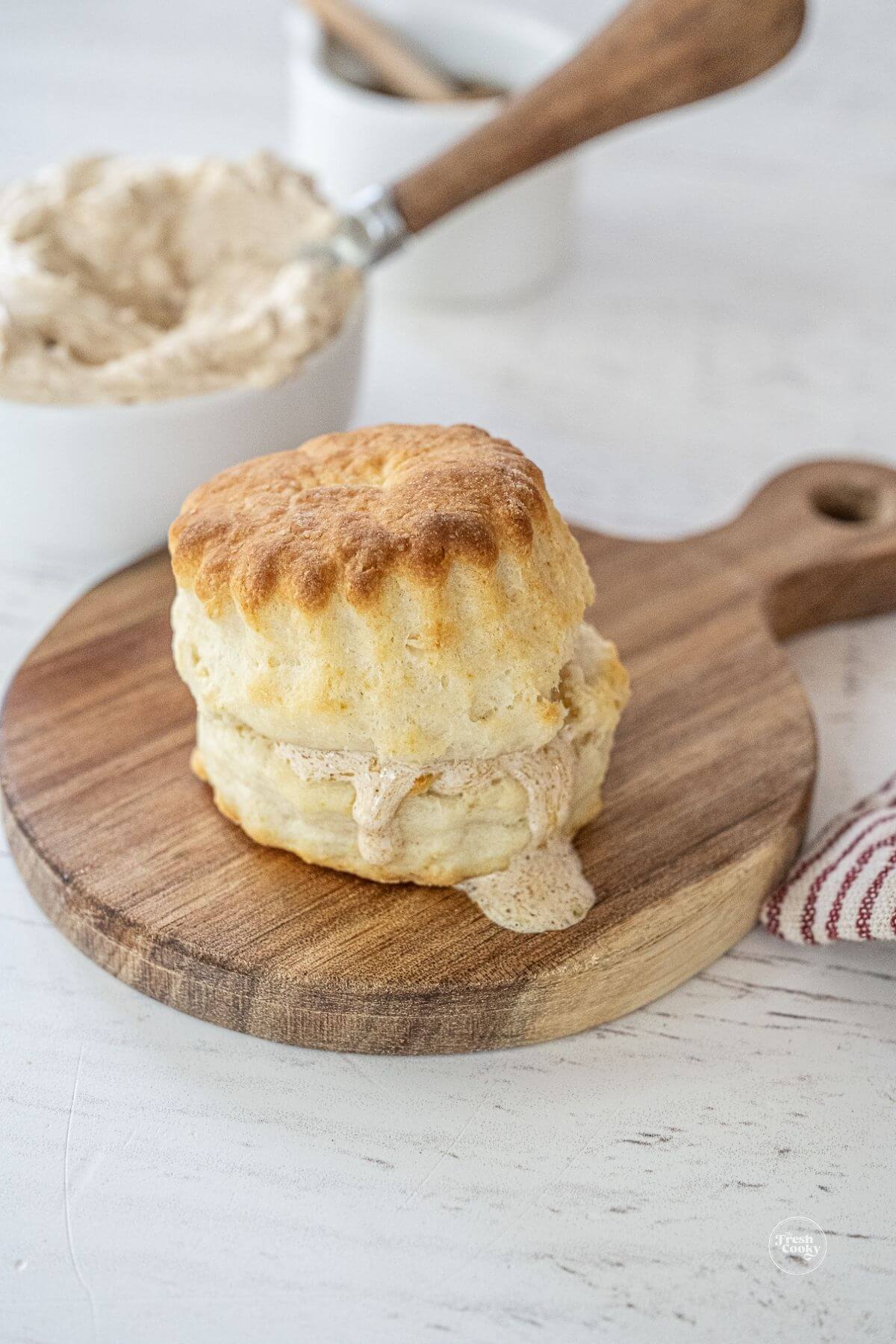 Hot biscuit with melting honey cinnamon butter on cutting board with bowl of honey butter in background.