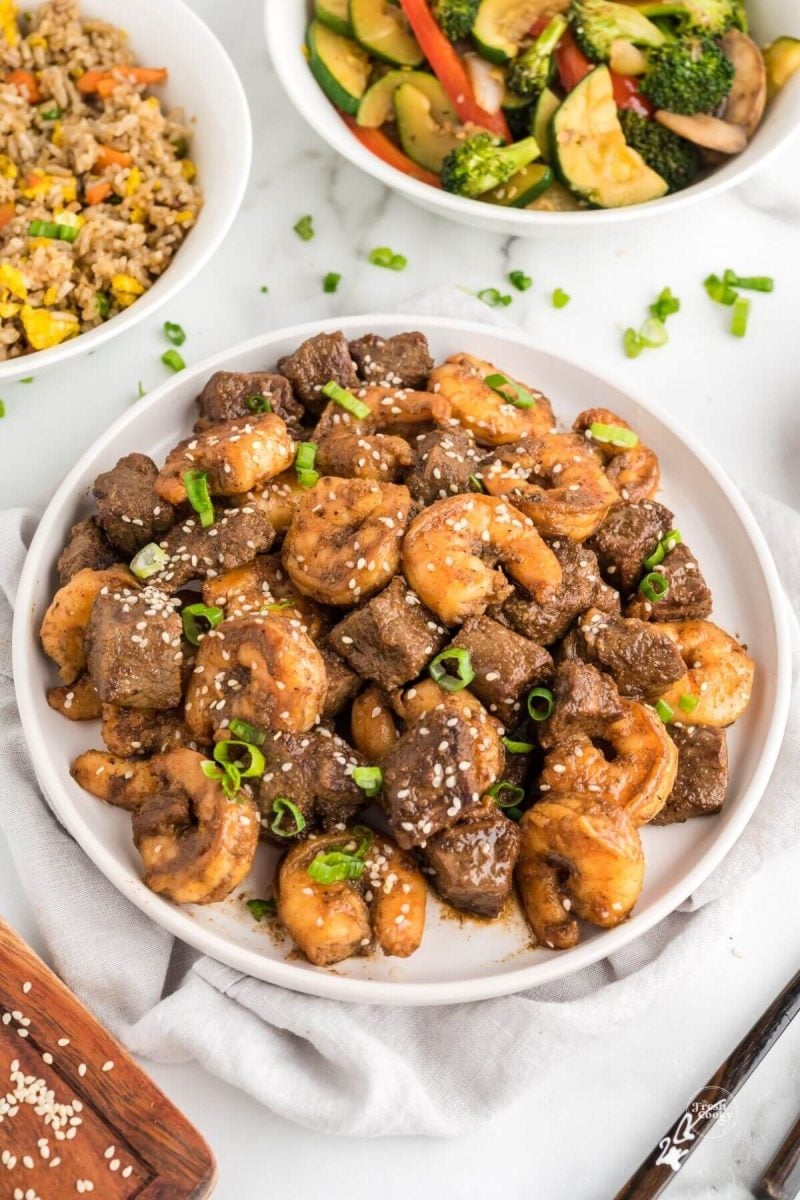 Steak and shrimp made on hibachi in serving bowl with fried rice and hibachi veggies in background. 