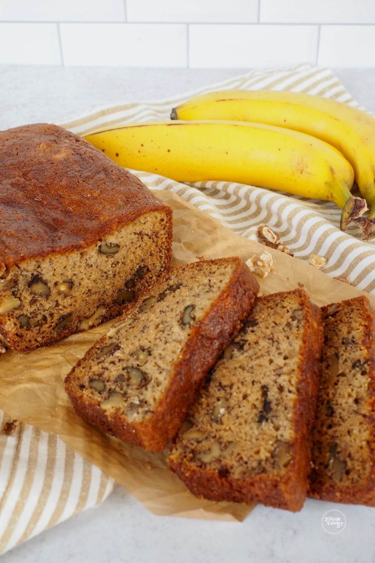 Slices on tray of high altitude banana bread with walnuts or chocolate chips with ripe bananas behind.