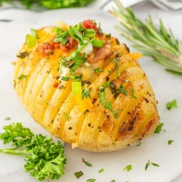 Garlic butter and herb Hasselback potato on plate topped with cheese, bacon bits and herbs.