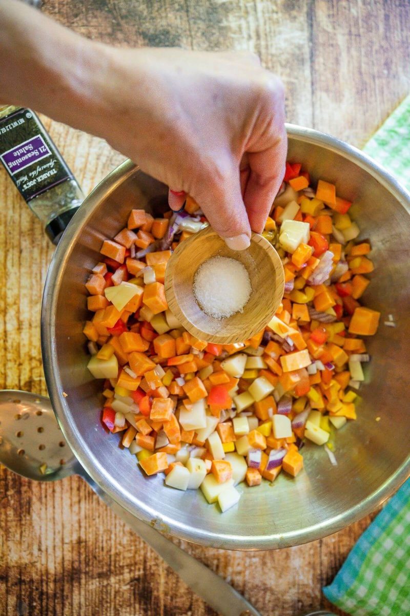 Adding kosher salt and pepper to bowl with diced sweet potatoes and vegetables for hash browns. 