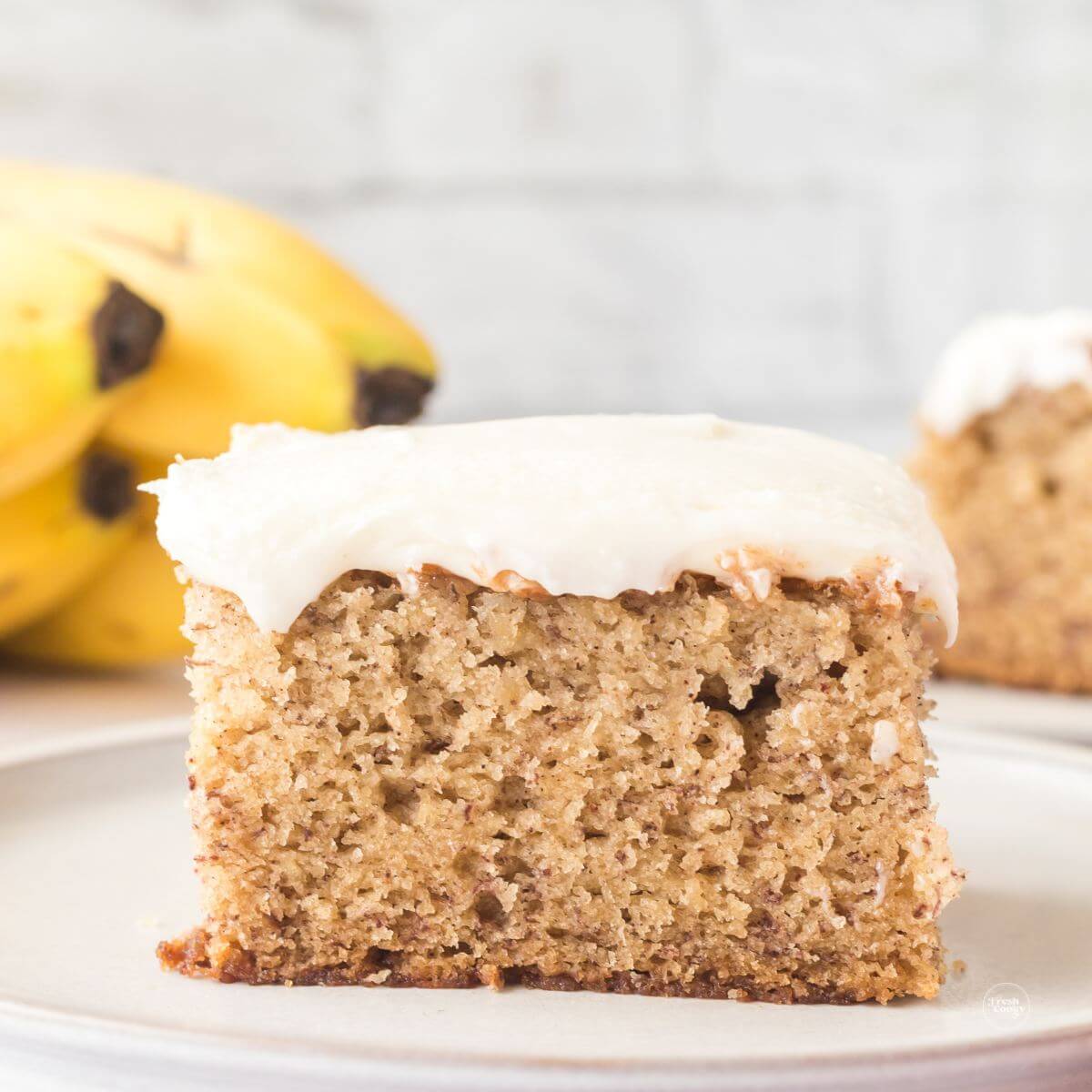 Square of banana bars topped with cream cheese frosting bananas in background.