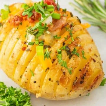 Garlic butter and herb Hasselback potato on plate topped with cheese, bacon bits and herbs.