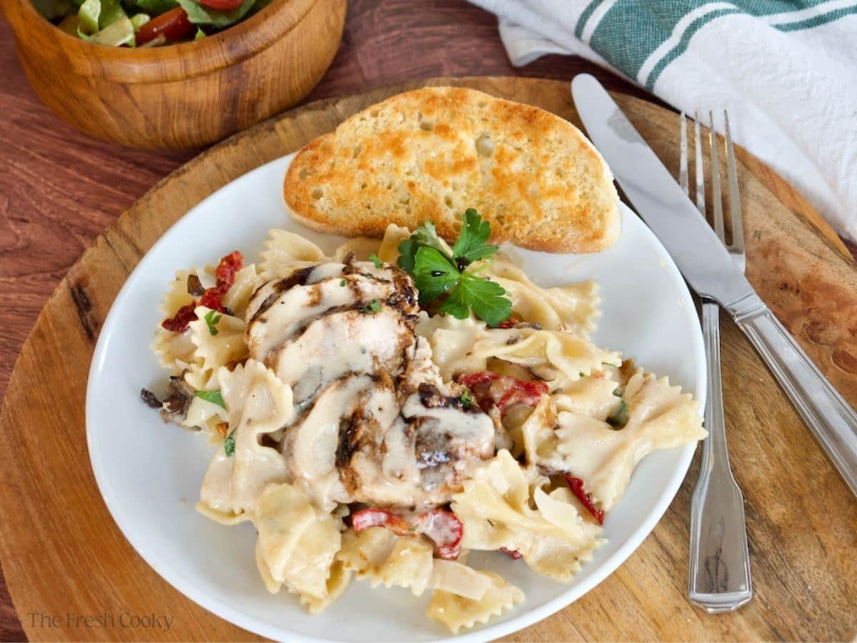 A white plate of creamy bow tie pasta topped with grilled chicken, a slice of garlic bread and a salad in the background.