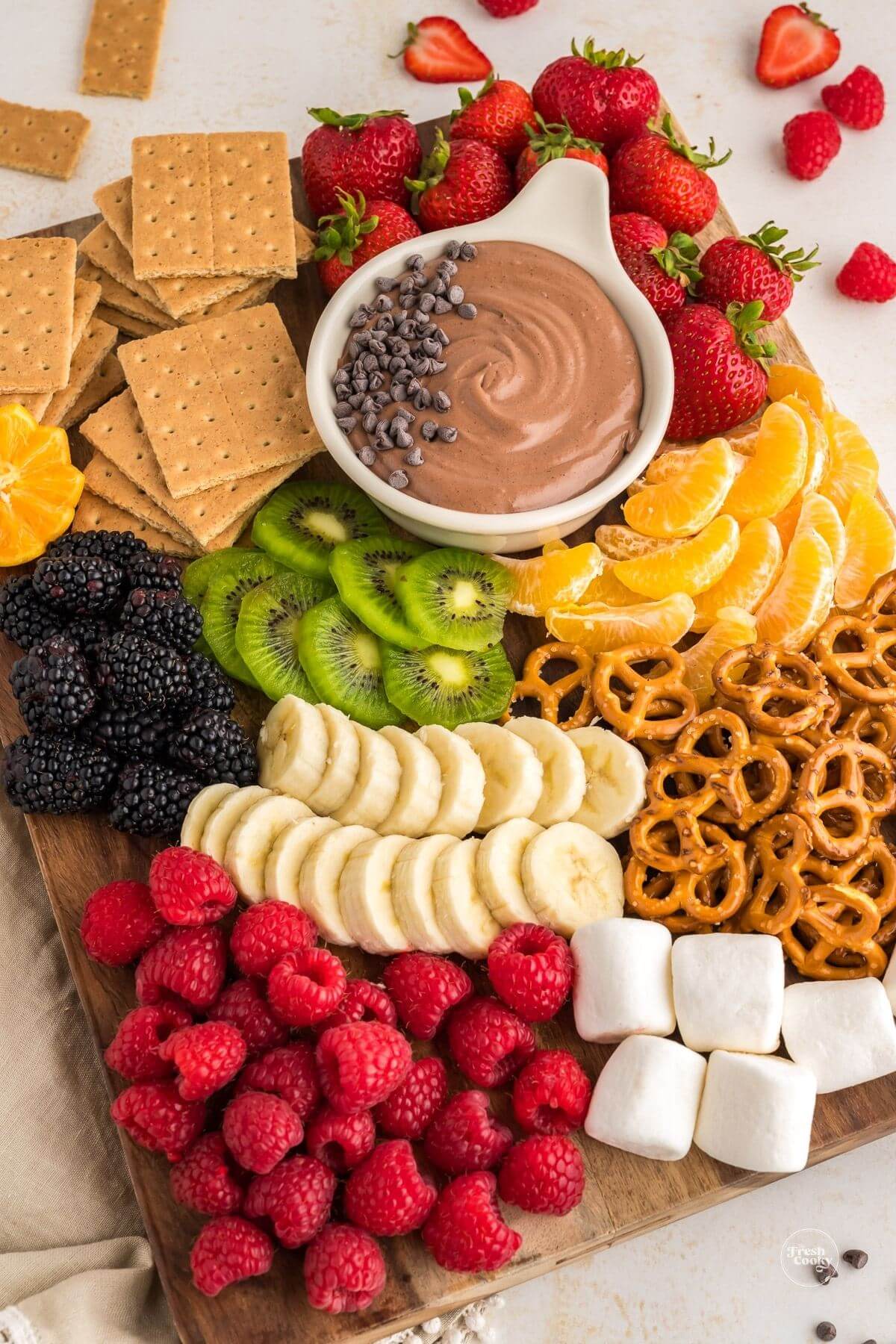 Chocolate fruit dip i bowl on board filled with fruits and graham crackers, and marshmallows.