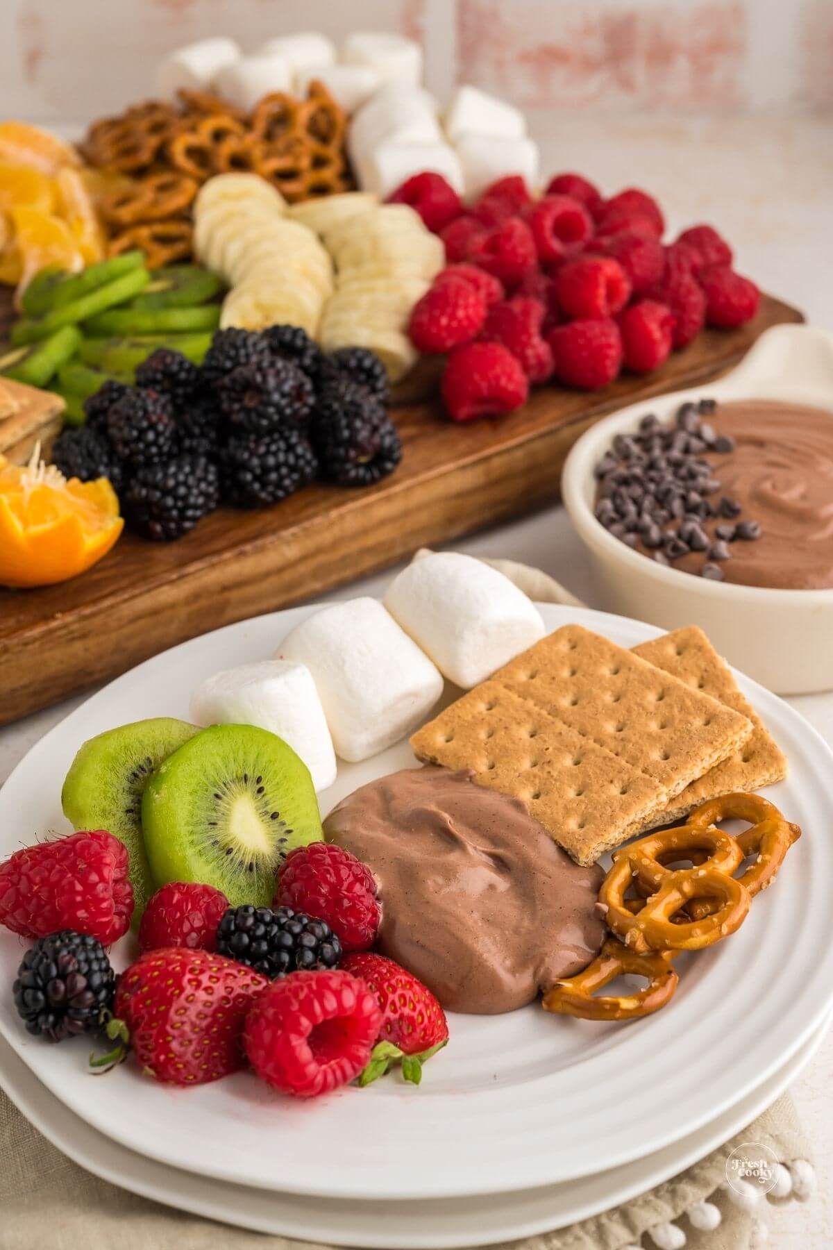 Chocolate dip serving on plate with assorted dippers and dip and assorted fruit and pretzels in background.