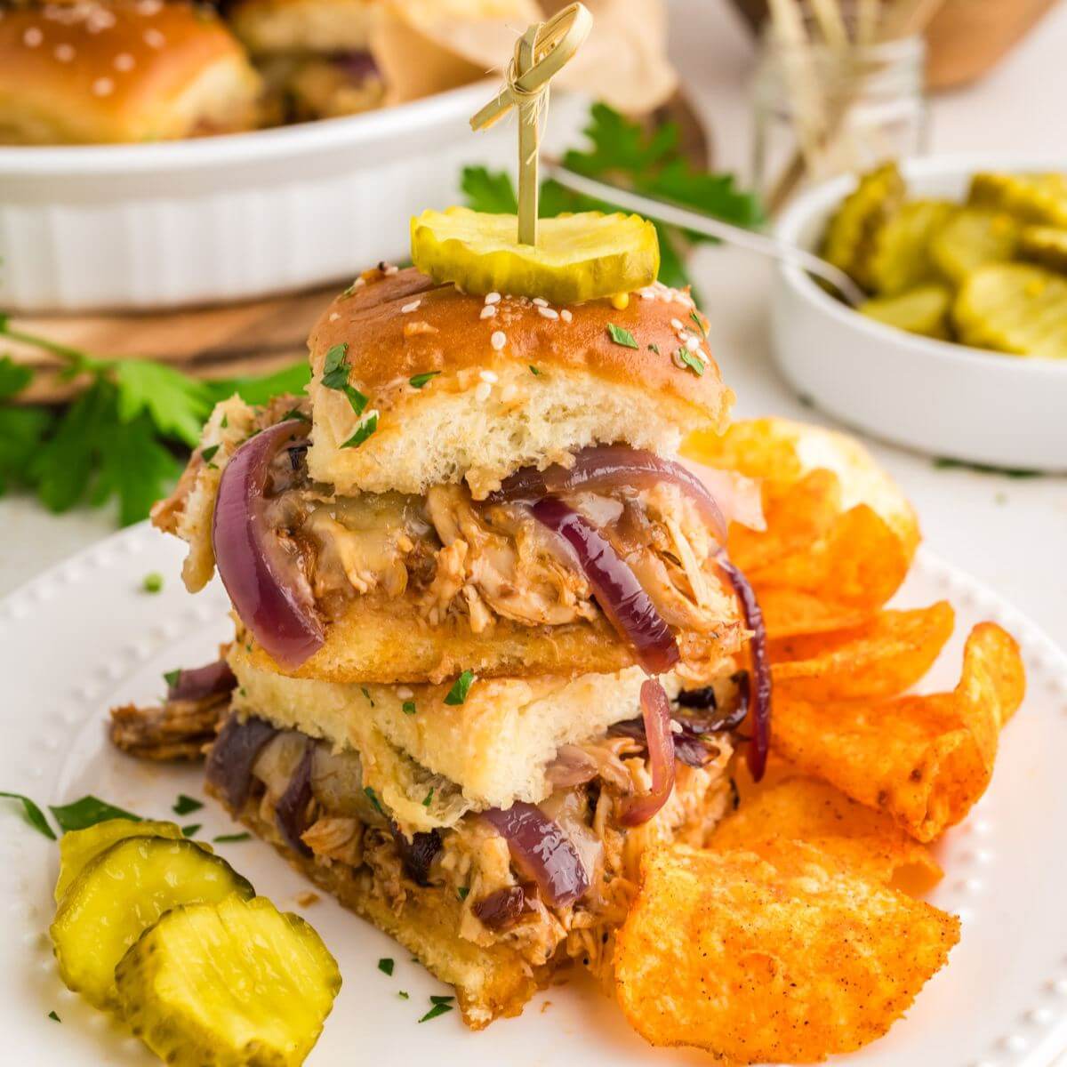 square image of stacked Barbeque Chicken Sliders topped with pickle and served with chips on plate.