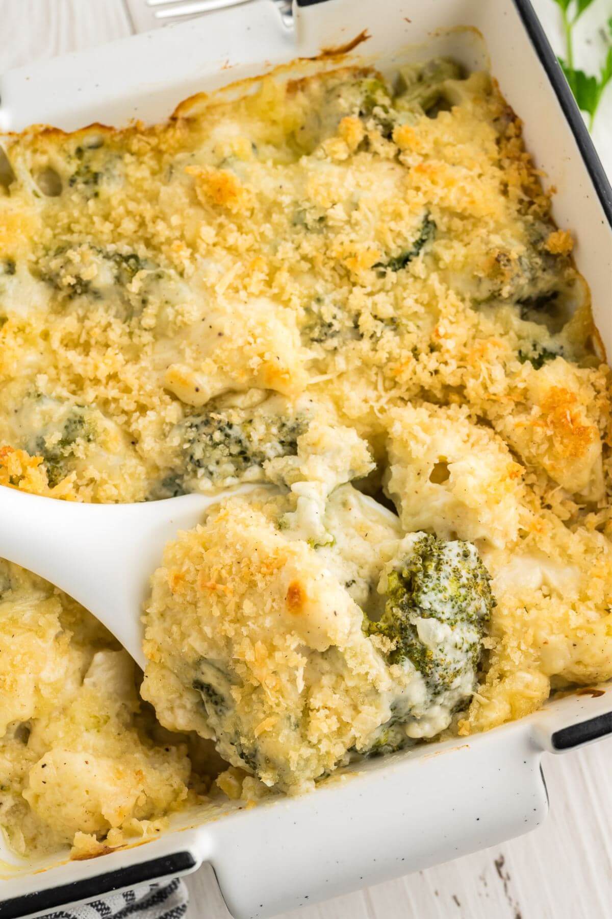 There is a spoon about to lift some Cheesy Broccoli Cauliflower Casserole out of the casserole dish.