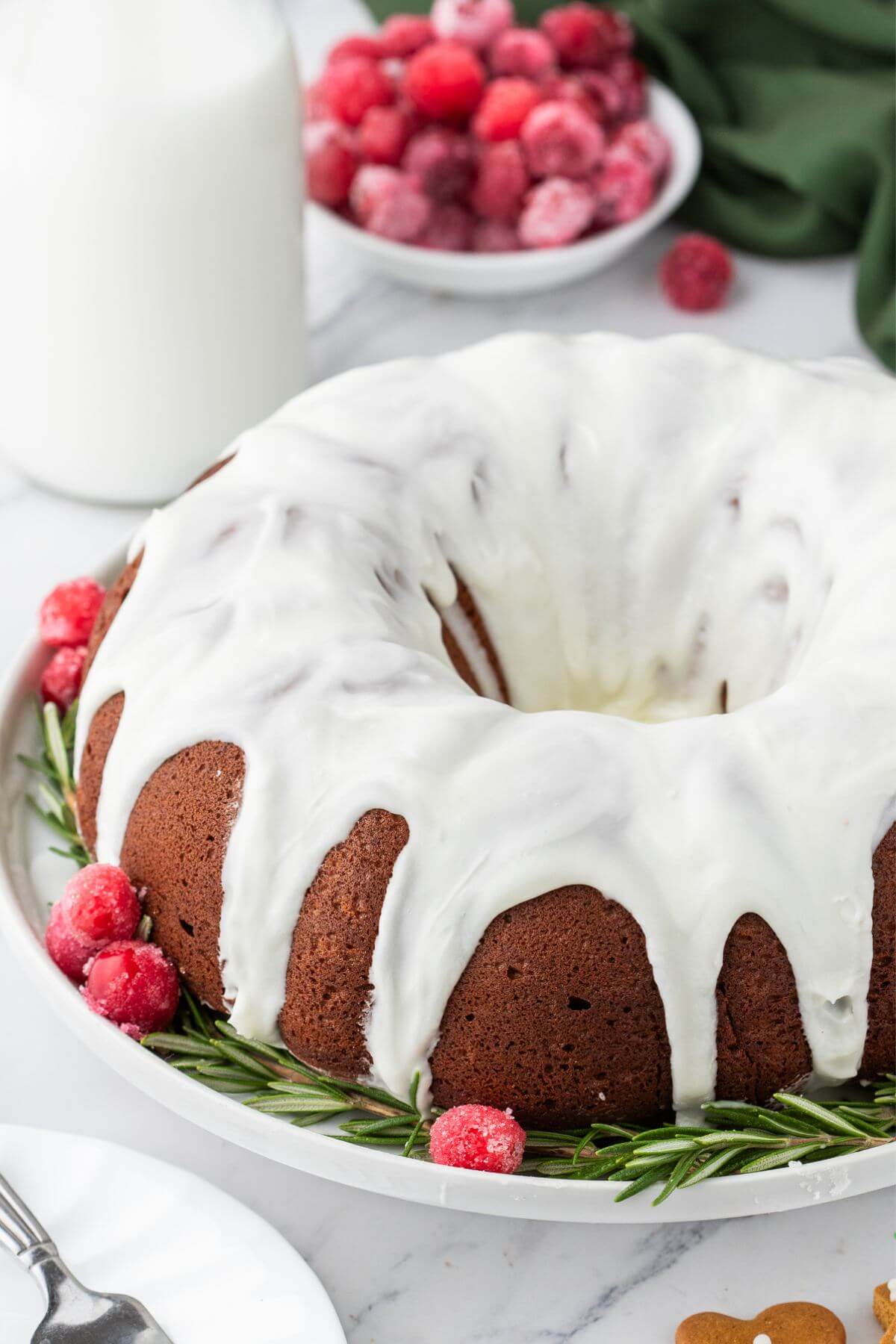 Whole gingerbread bundt cake iced with a bright white glaze and garnished with sugared cranberries and fresh rosemary.