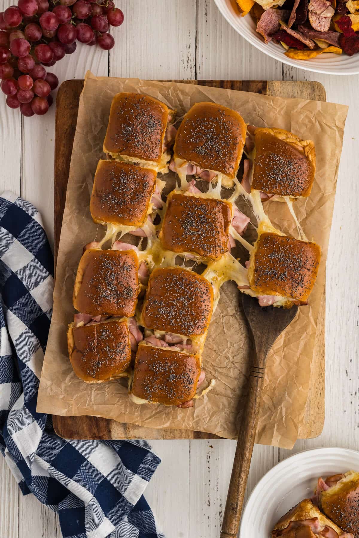 A bunch of ham and cheese sliders on a cutting board lined with parchment paper and pulling one out with a serving spatula.