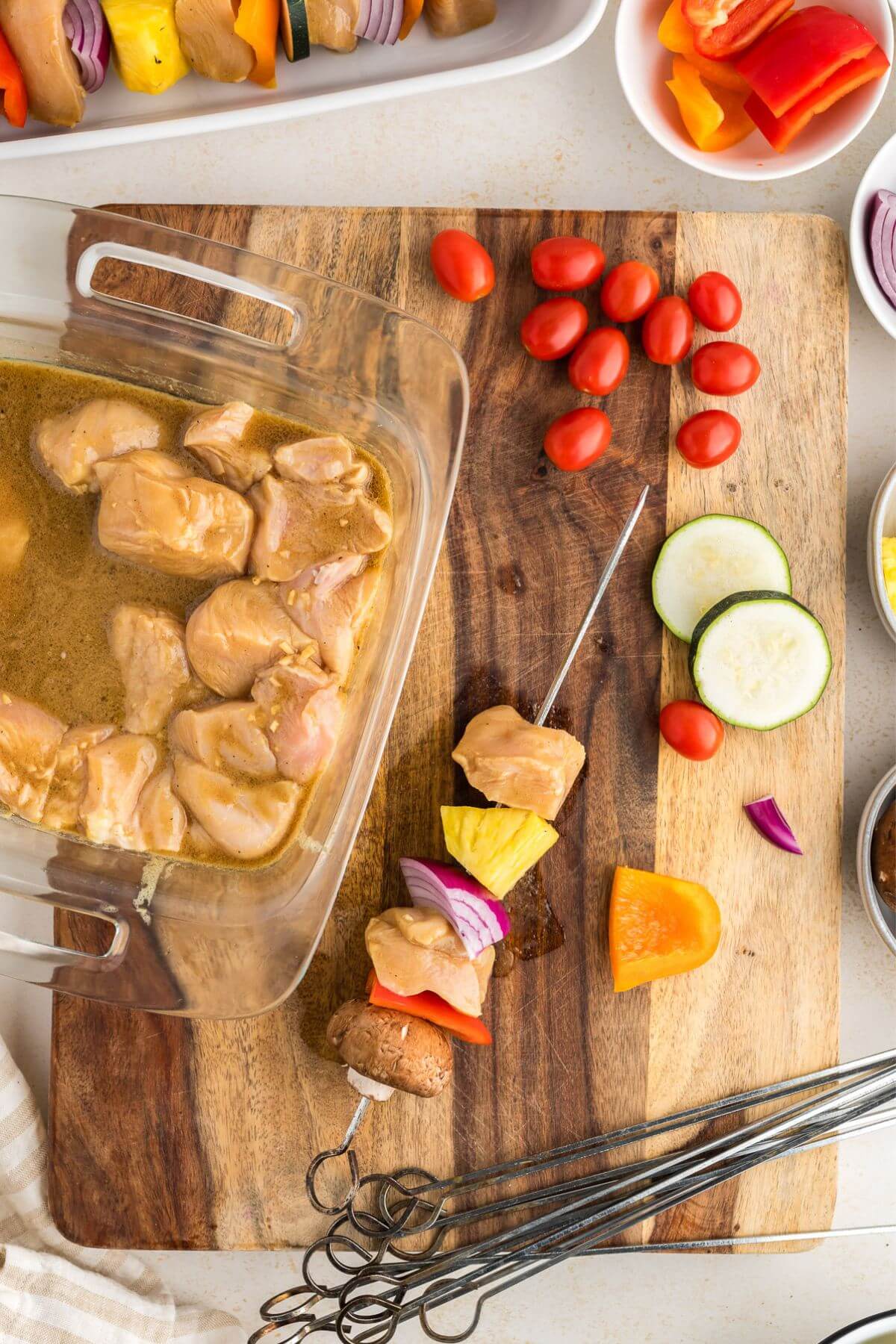 Cutting board with the dish of marinating chicken chunks and a skewer being assembled with veggies and meats near a group of skewers to make.
