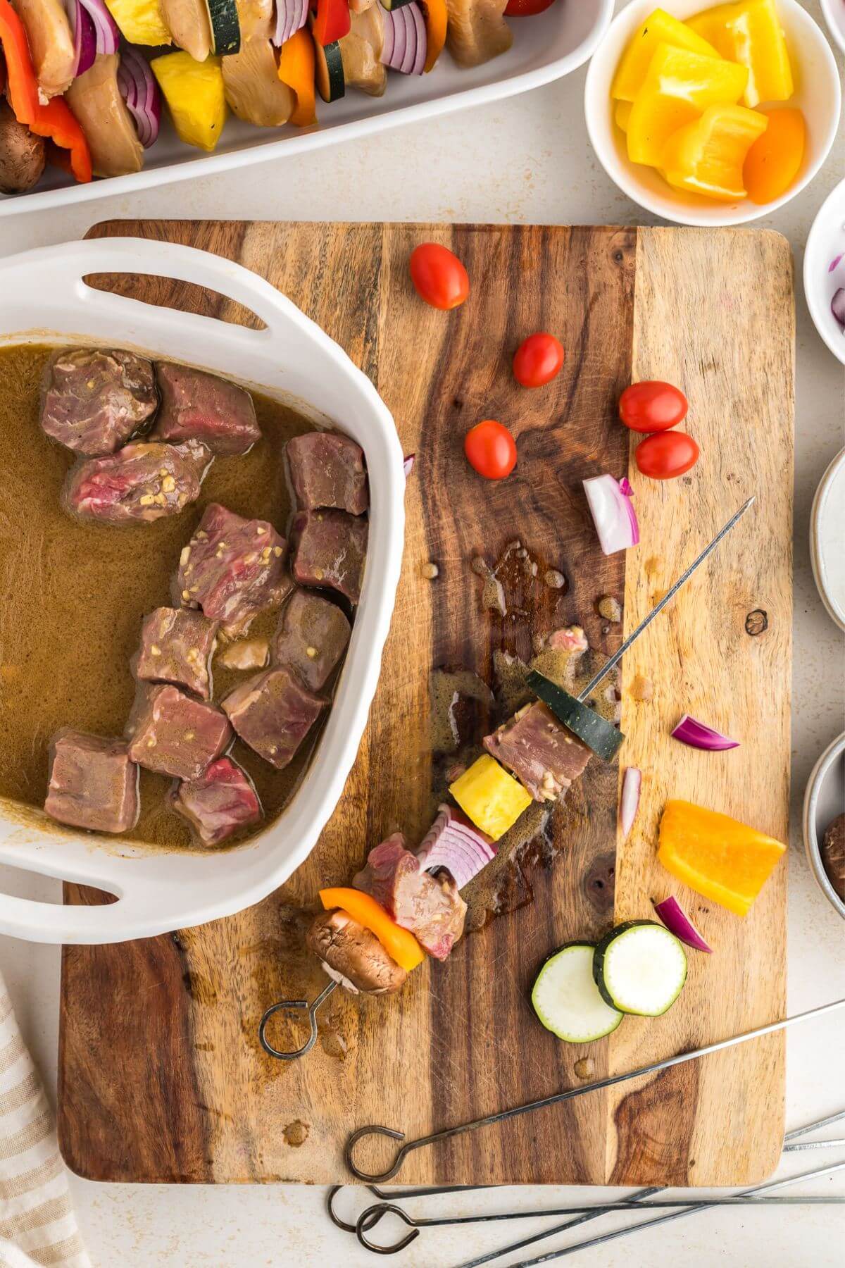 A cutting board with the dish of marinating steak chunks next to a skewer being assembled with veggies and steak.