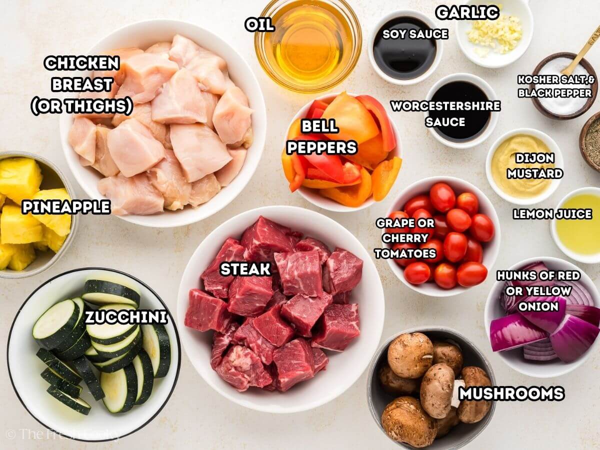 Labeled ingredients for shish kabobs. L-R, zucchini, pineapple, chicken breast chunks, oil, soy sauce, Worcestershire sauce, garlic, salt and pepper, dijon mustard, lemon juice, red onion chunks, button mushrooms, grape tomatoes and bell pepper chunks.
