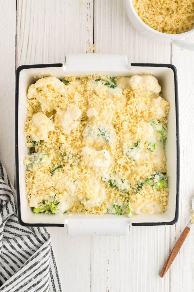 Cheese broccoli cauliflower casserole before baking, in casserole dish from the top next to fine napkin and bowl of panko breadcrumbs.