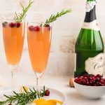 Two cranberry mimosas by champagne bottle and cranberries.