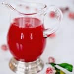 Cranberry simple syrup in glass pitcher surrounded by sugared cranberries and greens.