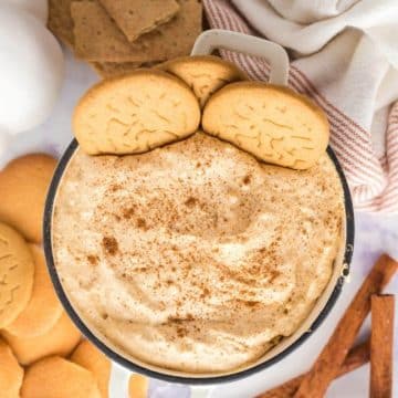 Pumpkin Fluff dip dessert with cookies artfully shown in dip and surrounding the bowl along with cinnamon sticks, graham crackers, and decorations.